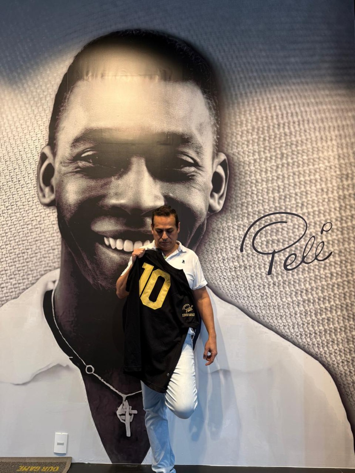 O artista apresenta camisa preta diante de retrato monumental de Pelé.